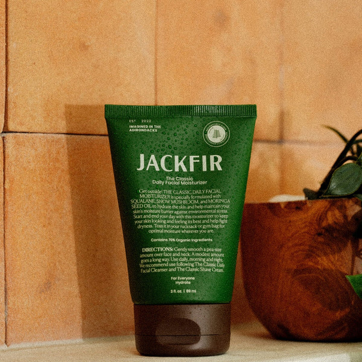 Jackfir Classic Daily Facial Moisturizer green tube standing upright with small potted plant beside it on sandy surface