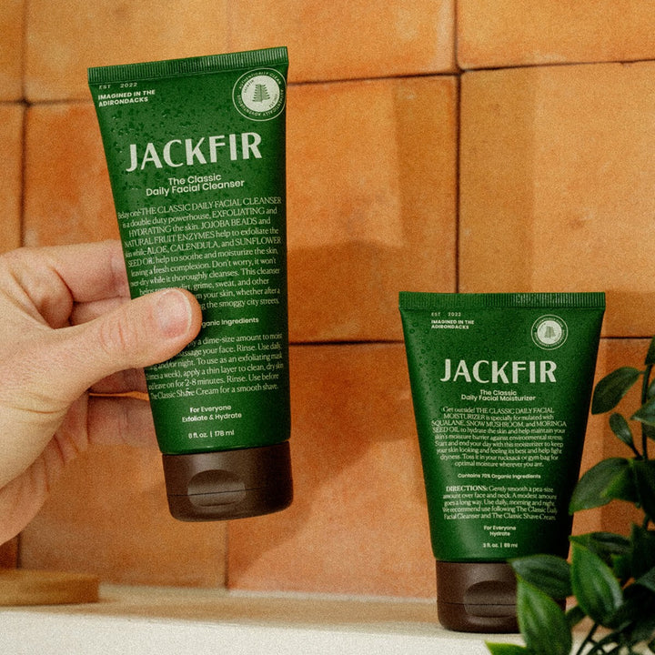 Hand holding Jackfir Cleanser tube with Moisturizer standing beside it on orange tile with green plant