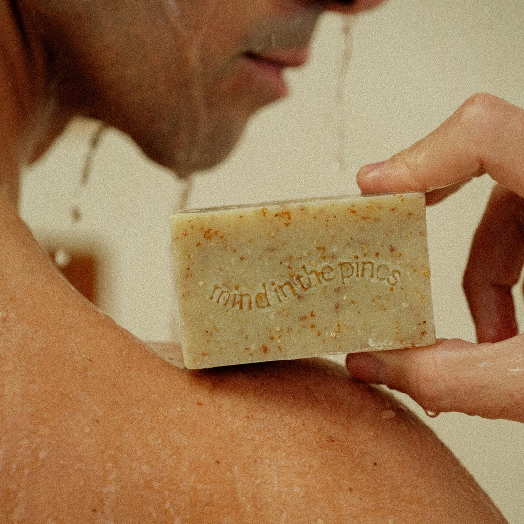 Hands holding single Jackfir The Bushwhacker Bar Soap showing wheat bran specks and embossed text near bare shoulder