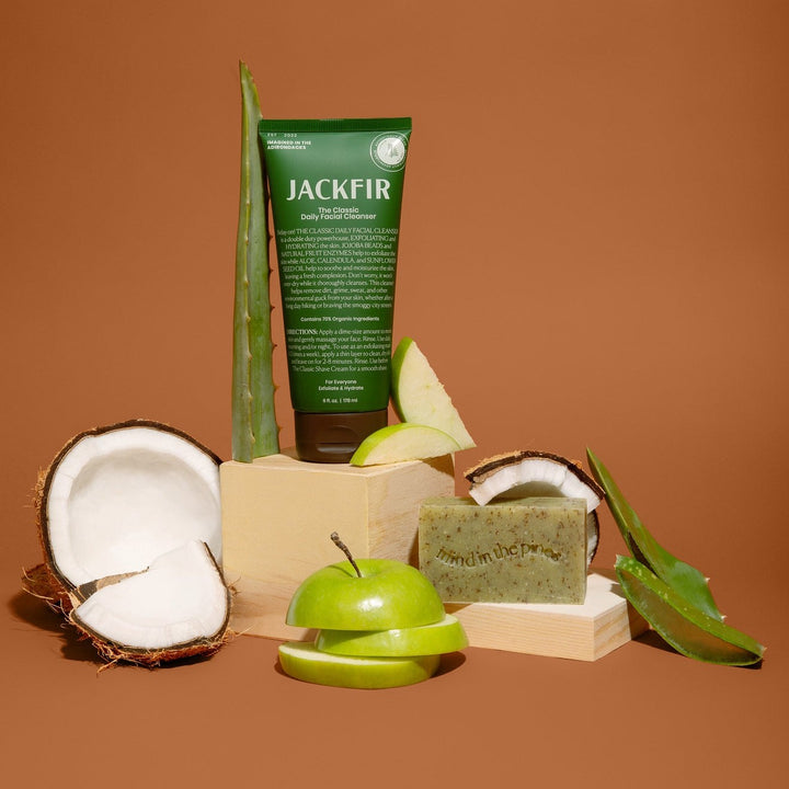 Jackfir The Exfoliating Duo green tube and speckled bar soap on wood blocks with coconut, apple, and aloe props